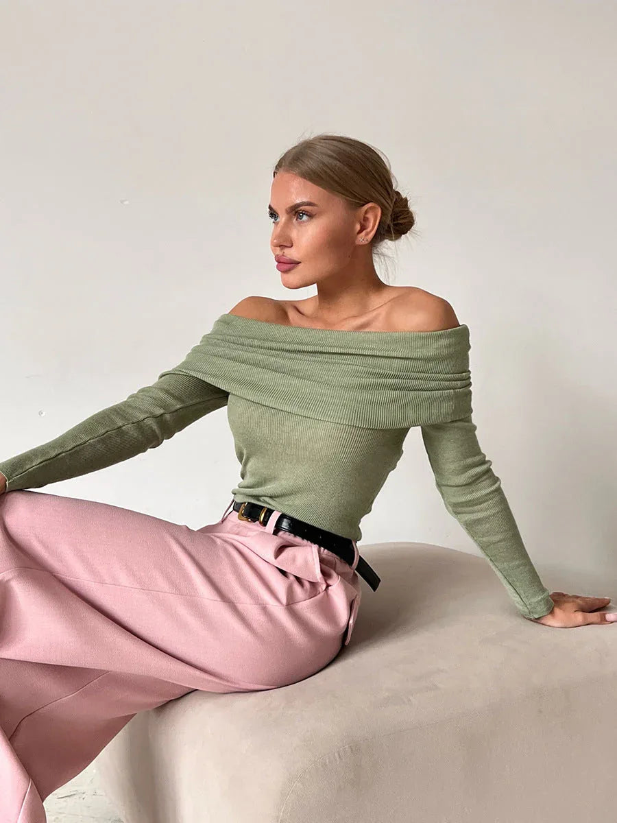 Woman wearing a green off-the-shoulder ribbed knit sweater with long sleeves and pink wide-leg pants, sitting on a light-colored surface.