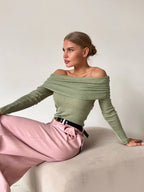 Woman wearing a green off-the-shoulder ribbed knit sweater with long sleeves and pink wide-leg pants, sitting on a light-colored surface.