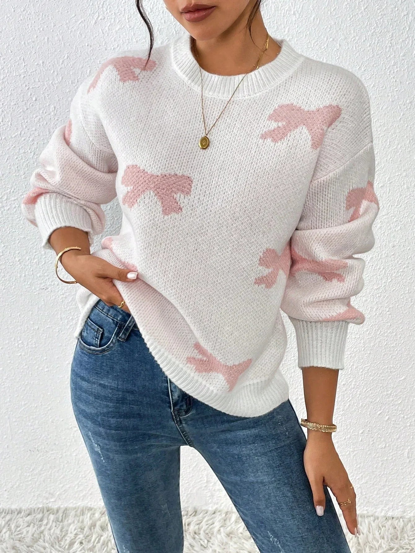 Woman wearing a white crew neck sweater with pink bow print and blue jeans, holding her hand on her hip