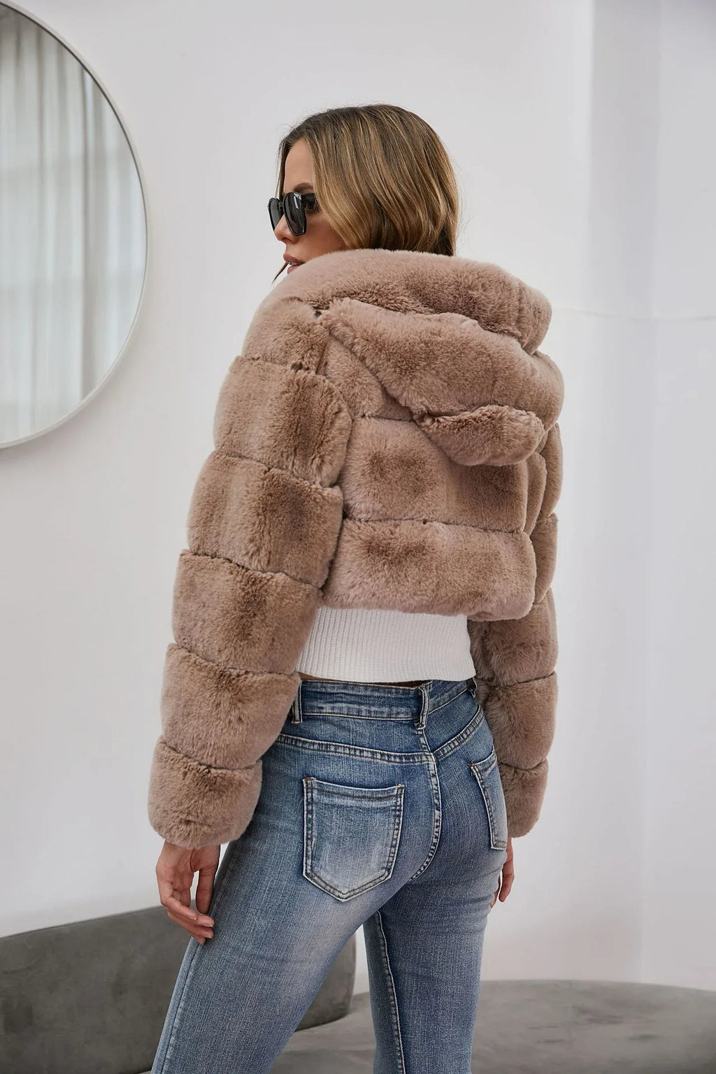 Woman wearing a cropped faux fur hooded jacket over a white camisole and blue jeans, back view.