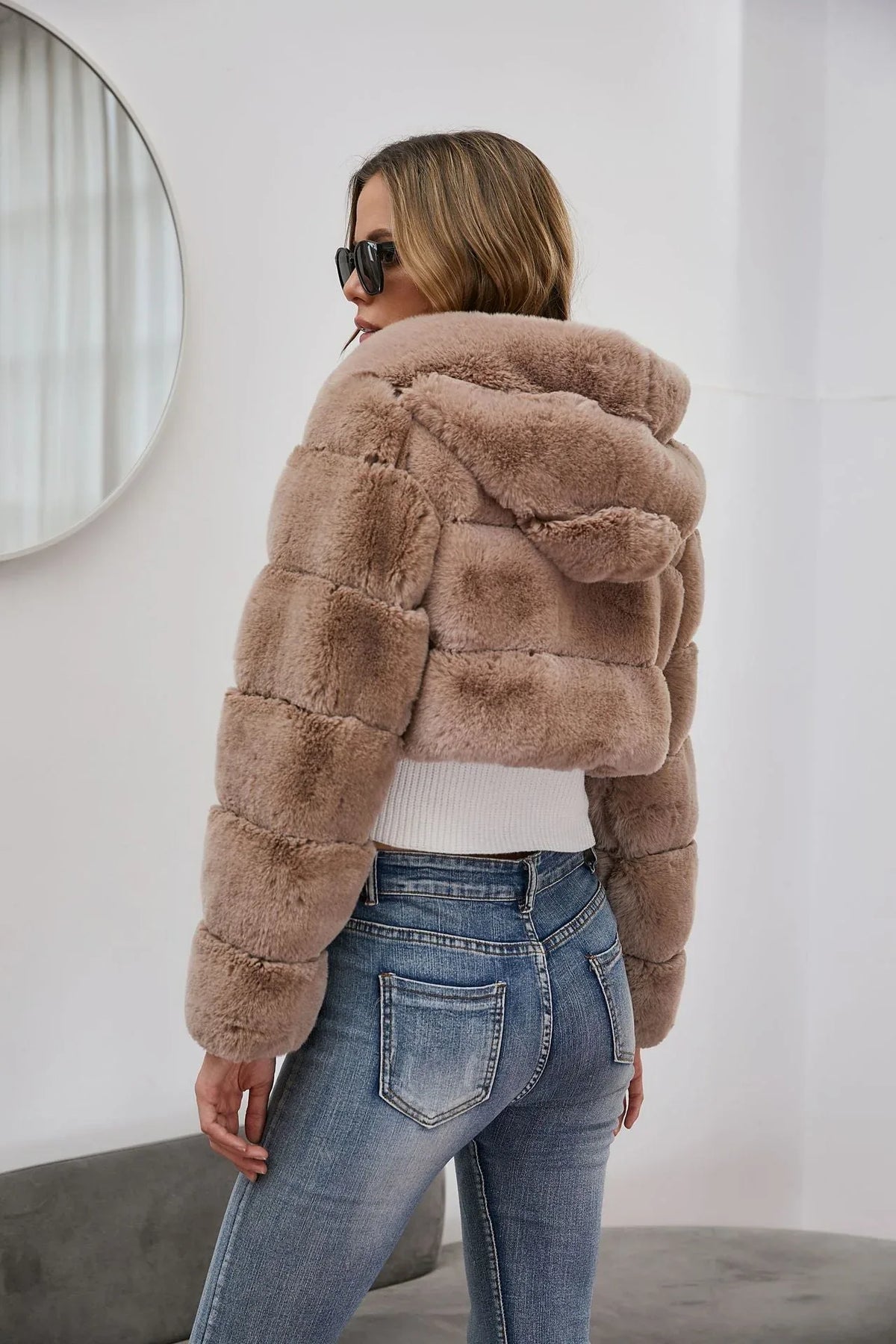 Woman wearing a cropped faux fur hooded jacket over a white camisole and blue jeans, back view.