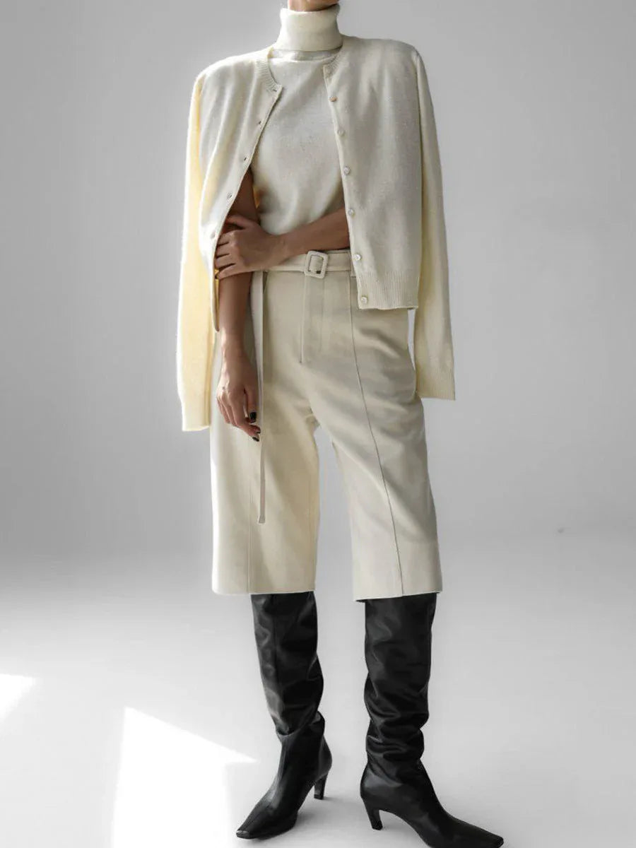 Full body shot of a woman wearing a cream-colored cashmere cardigan over a matching turtleneck, cream-colored culottes, and black knee-high boots.
