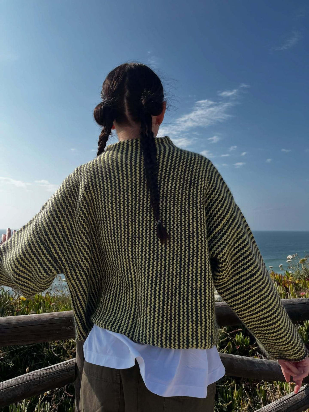 Back view of a woman wearing a green and black striped knit cardigan with colorful buttons, arms outstretched, by the ocean.