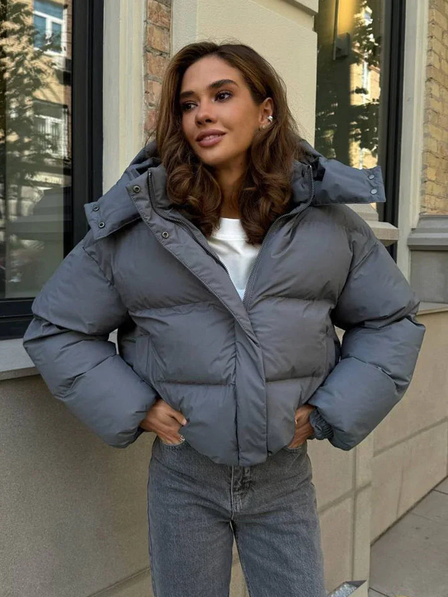 Woman wearing a grey cropped puffer jacket with a hood, white t-shirt, and grey jeans, standing outdoors.