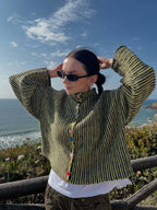 Woman wearing a green and black striped knit cardigan with colorful buttons, adjusting her hair, by the ocean.