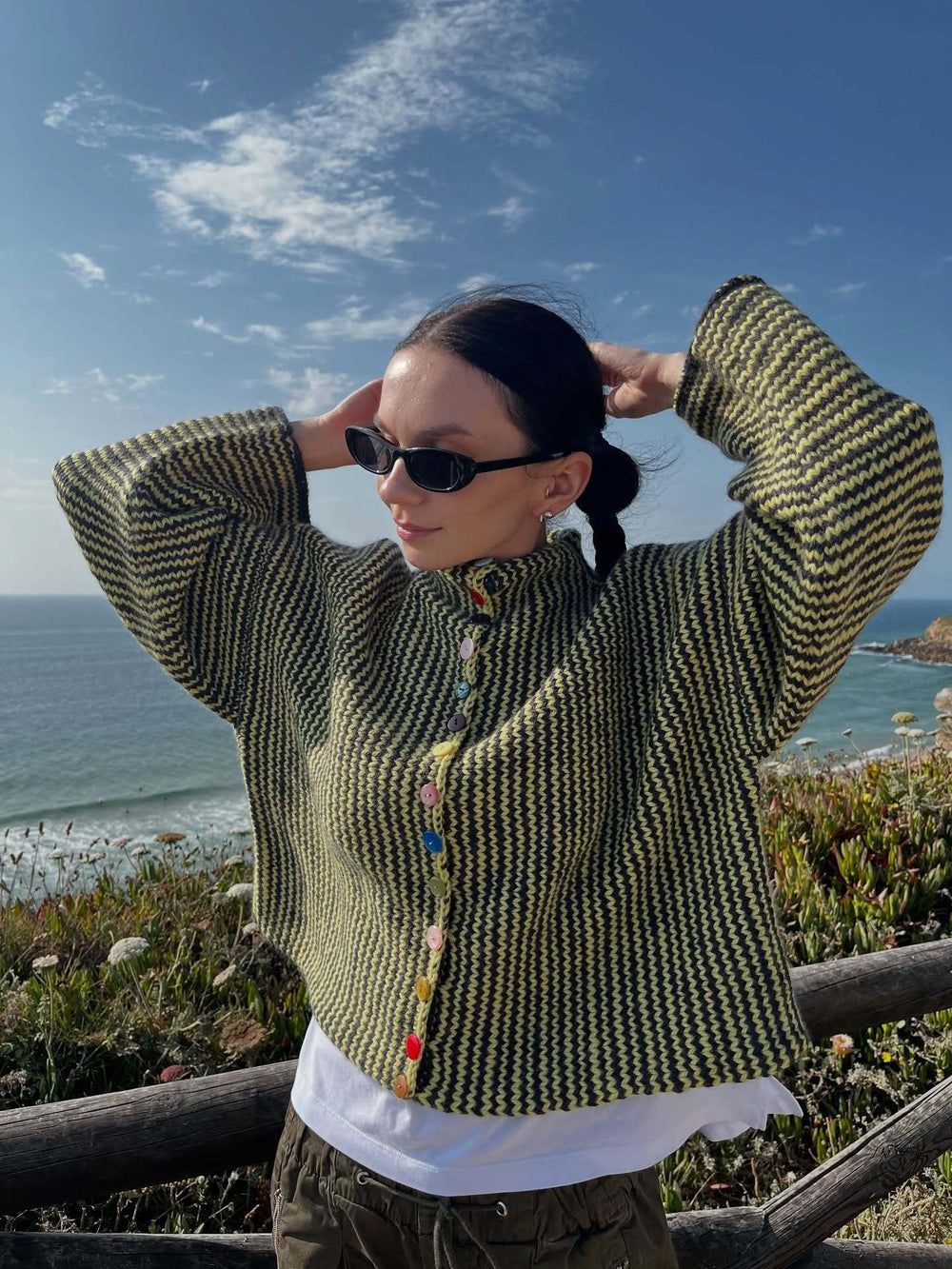 Woman wearing a green and black striped knit cardigan with colorful buttons, adjusting her hair, by the ocean.