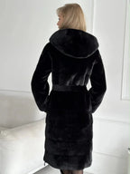 Back view of a woman wearing a black hooded faux fur midi coat with a belted waist.