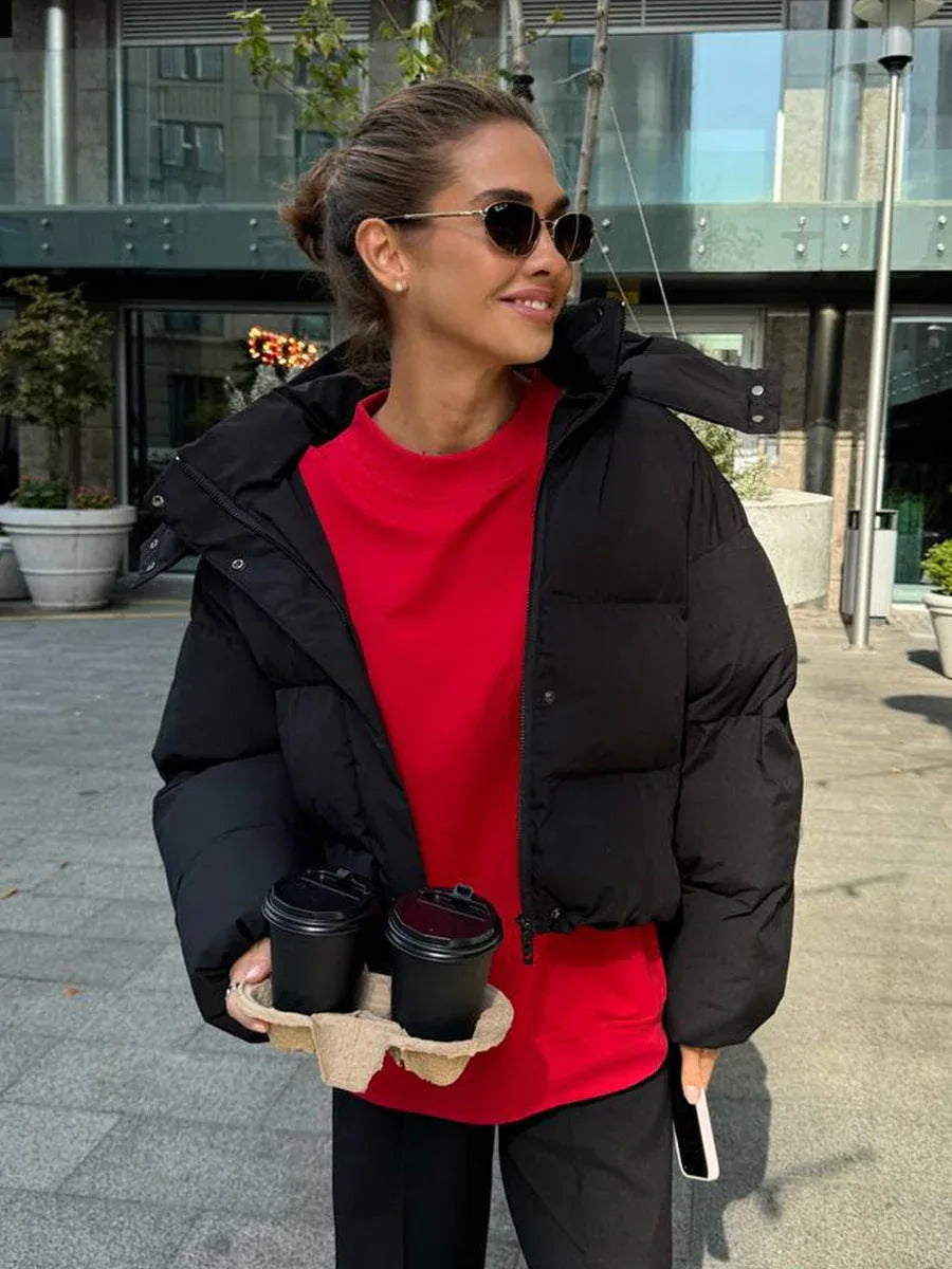 Woman wearing a black cropped puffer jacket with a hood, red t-shirt, and black pants, holding two coffee cups outdoors.