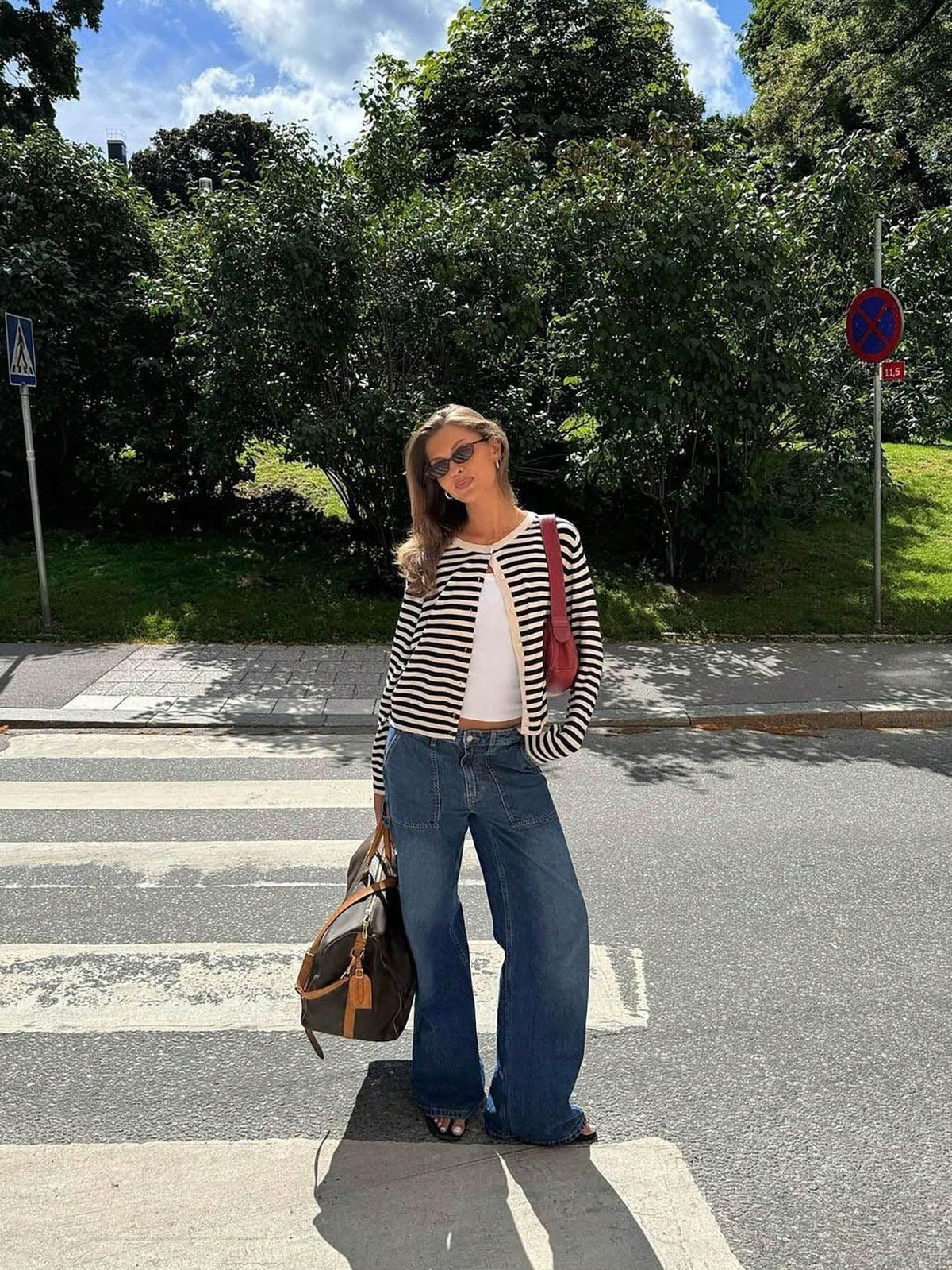 Full-body shot of a woman in a black and white striped cardigan, white tank top, and wide-leg jeans, standing on a crosswalk with a duffel bag and red purse.
