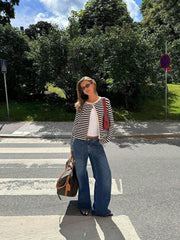 Full-body shot of a woman in a black and white striped cardigan, white tank top, and wide-leg jeans, standing on a crosswalk with a duffel bag and red purse.