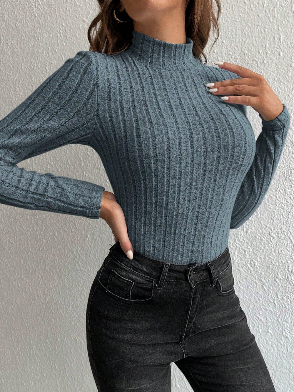 Close-up front view of a woman wearing a fitted, long-sleeved, ribbed mock neck top in a blue-grey color with black jeans, with hands placed on her chest and hip.