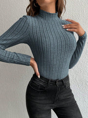 Close-up front view of a woman wearing a fitted, long-sleeved, ribbed mock neck top in a blue-grey color with black jeans, with hands placed on her chest and hip.