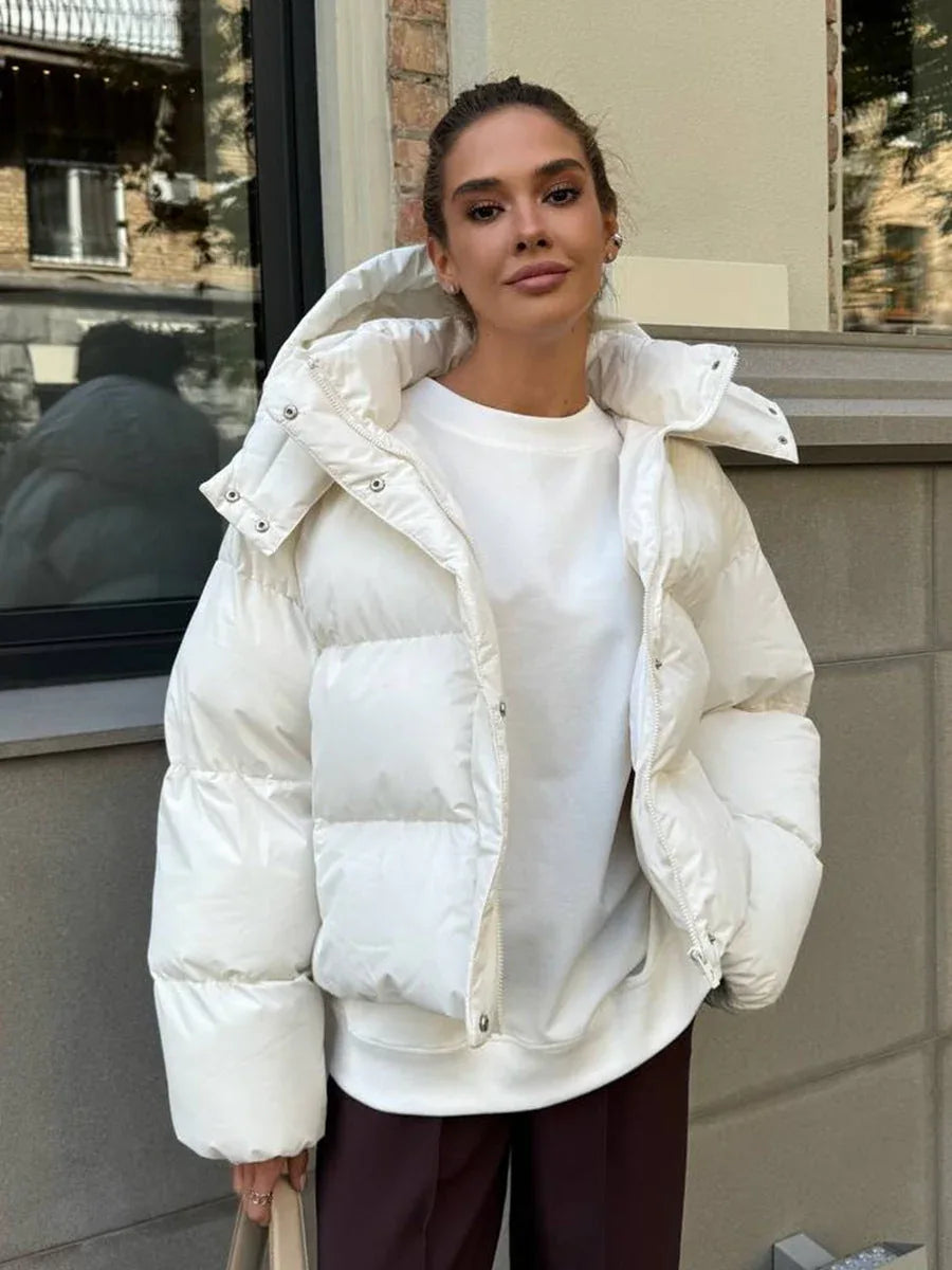 Woman wearing a white cropped puffer jacket with a hood, white t-shirt, and brown pants, standing outdoors. Main image