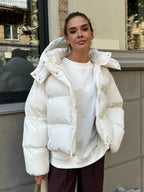 Woman wearing a white cropped puffer jacket with a hood, white t-shirt, and brown pants, standing outdoors.