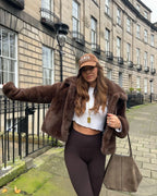 Woman wearing a brown faux fur cropped jacket, white crop top, and brown leggings, leaning against a railing.