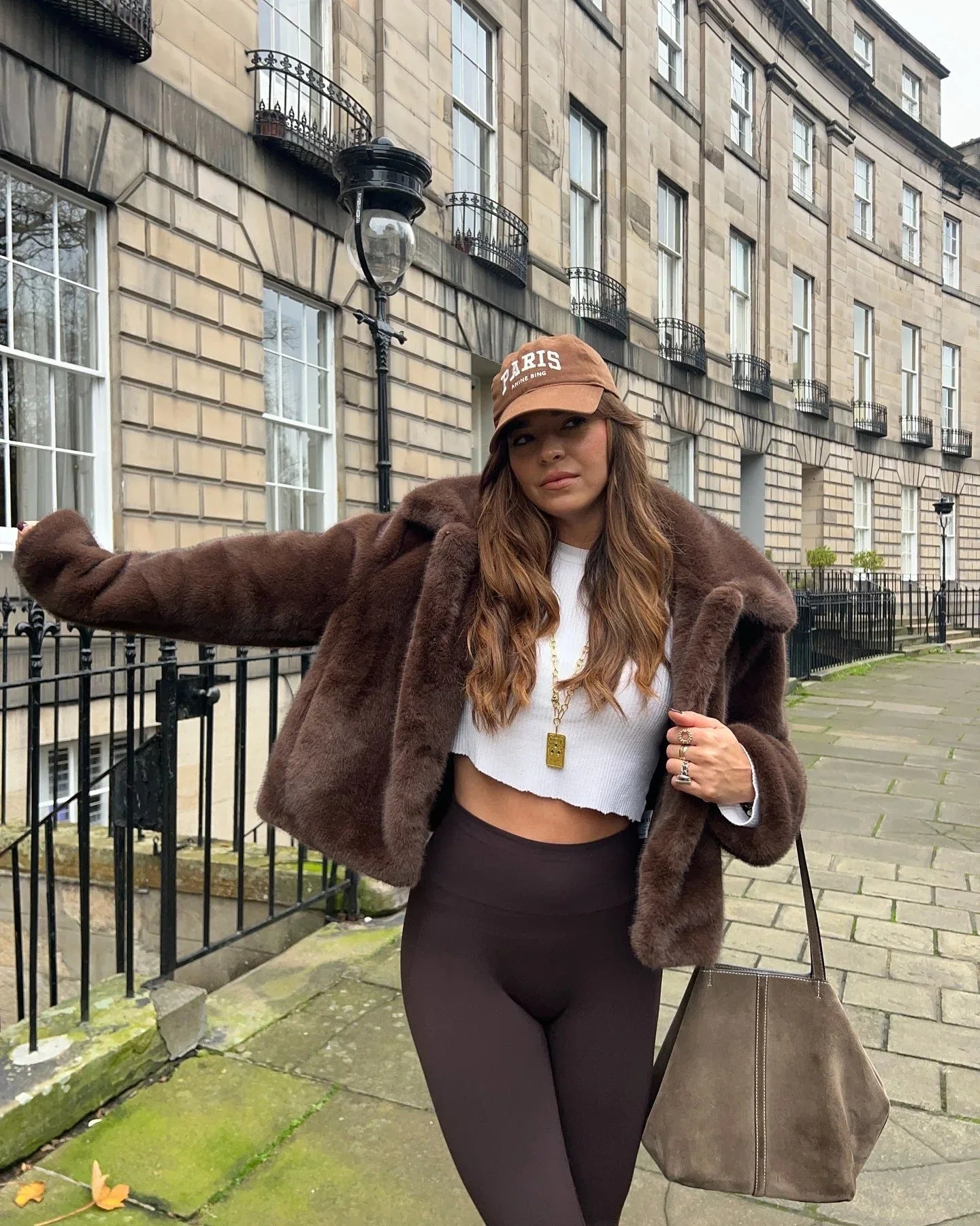 Woman wearing a brown faux fur cropped jacket, white crop top, and brown leggings, leaning against a railing.