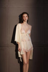 Woman standing, wearing a cream satin camisole and shorts set with pink lace trim, and a matching cream satin robe.