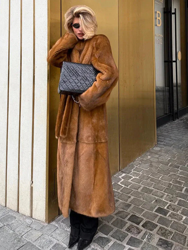 Woman posing in a long brown faux fur coat and black pants, holding a quilted black handbag.