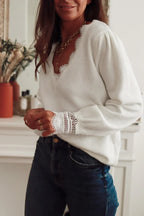 Woman wearing a white knit sweater with lace trim and blue jeans, hands clasped in front.