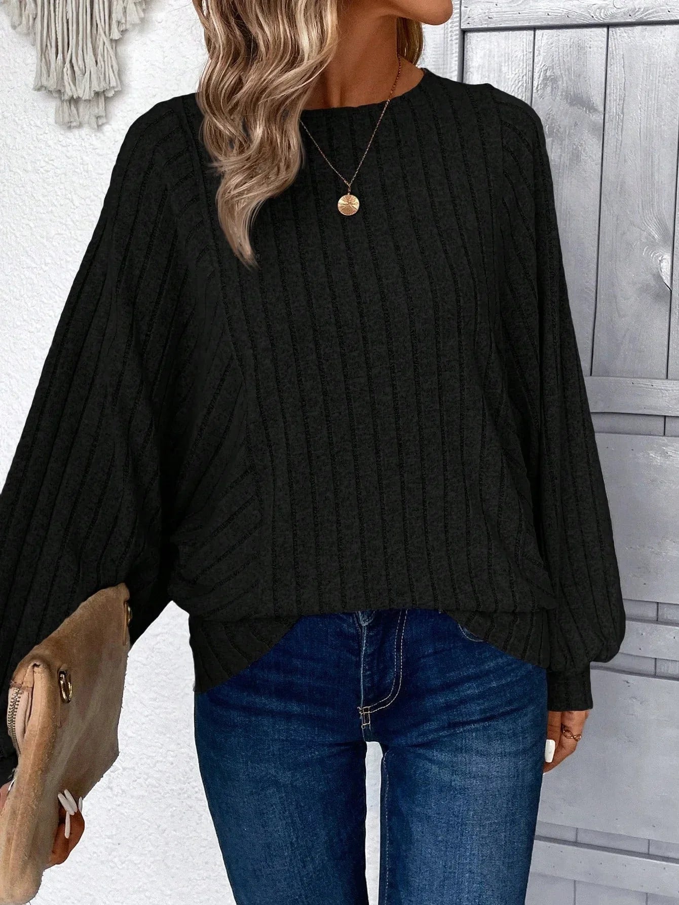 Woman wearing a black ribbed knit sweater with batwing sleeves and blue jeans, holding a tan clutch.