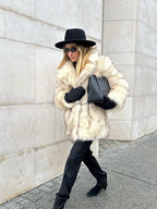 Woman in a cream faux fur coat, black hat, and black pants walking on a sidewalk.