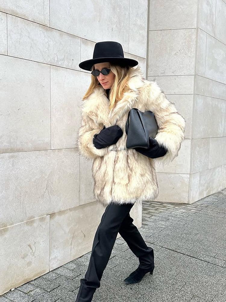 Woman in a cream faux fur coat, black hat, and black pants walking on a sidewalk.