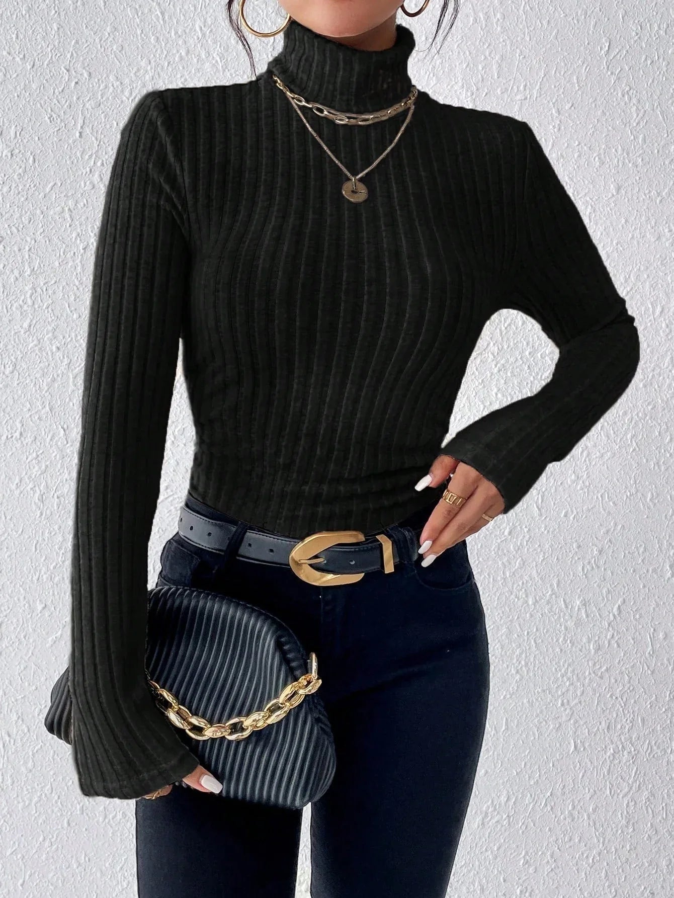 Woman wearing a black ribbed turtleneck sweater, black jeans, a black belt with a gold buckle, and holding a black clutch with a gold chain.
