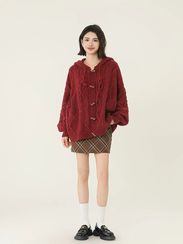 Woman standing with hands in pockets wearing a burgundy cable knit hooded cardigan and plaid mini skirt.