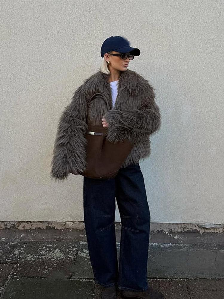 Woman standing and holding a brown tote bag, wearing a shaggy grey faux fur jacket, dark wash wide-leg jeans, and a navy baseball cap.