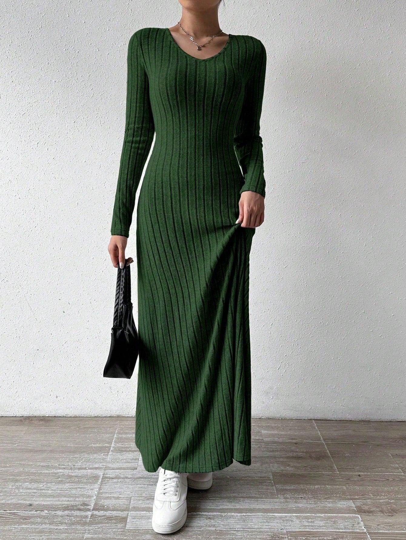 Woman wearing a long-sleeved green ribbed maxi dress, holding the hem of the dress and a black handbag, wearing white sneakers, posing against a white wall.