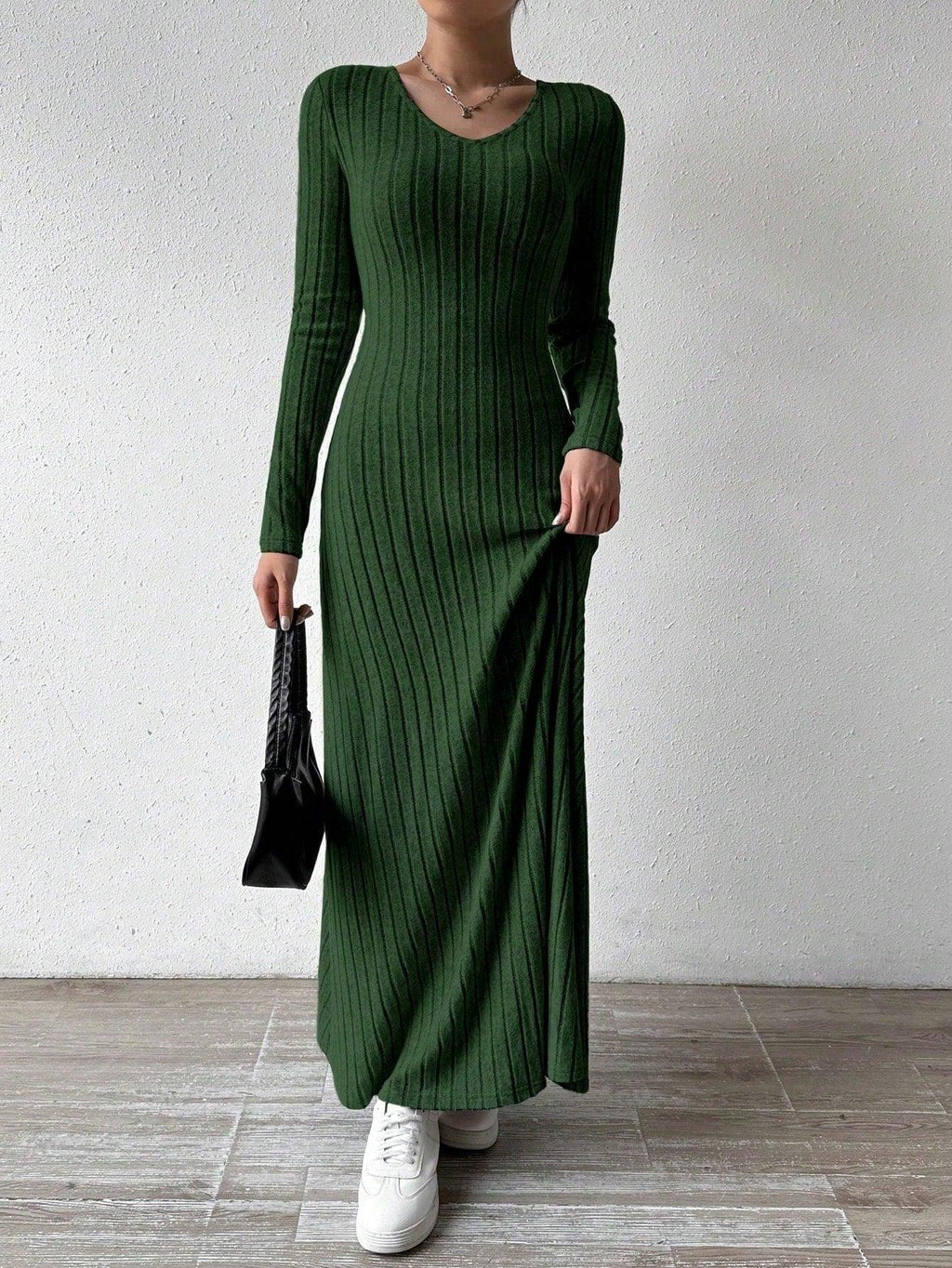 Woman wearing a long-sleeved green ribbed maxi dress, holding the hem of the dress and a black handbag, wearing white sneakers, posing against a white wall.