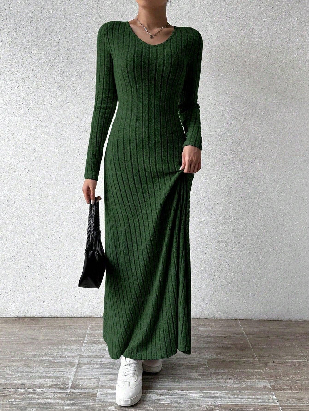 Woman wearing a long-sleeved green ribbed maxi dress, holding the hem of the dress and a black handbag, wearing white sneakers, posing against a white wall.