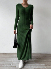 Woman wearing a long-sleeved green ribbed maxi dress, holding the hem of the dress and a black handbag, wearing white sneakers, posing against a white wall.