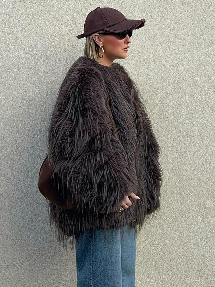 Woman in a brown shaggy faux fur jacket, blue jeans, and a brown baseball cap, viewed from the side.