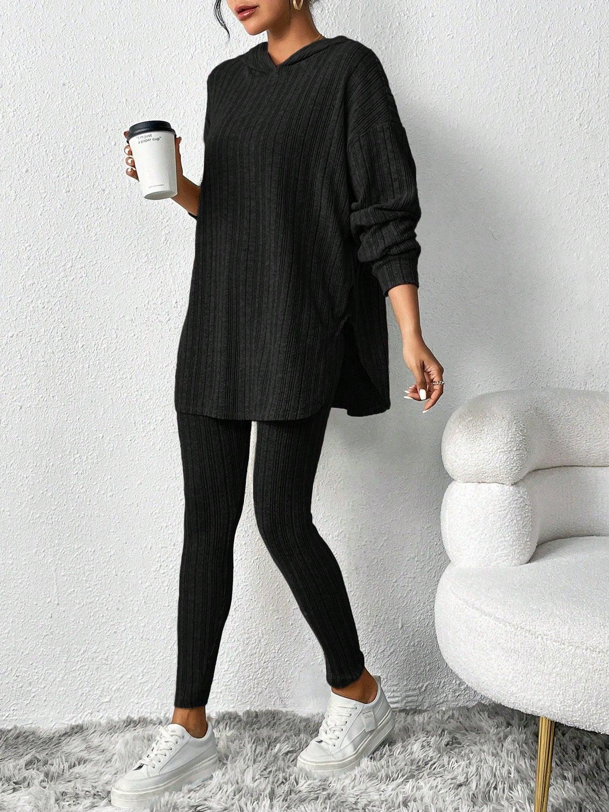 Woman wearing a black ribbed knit loungewear set consisting of a hooded tunic and leggings, holding a coffee cup. Main image