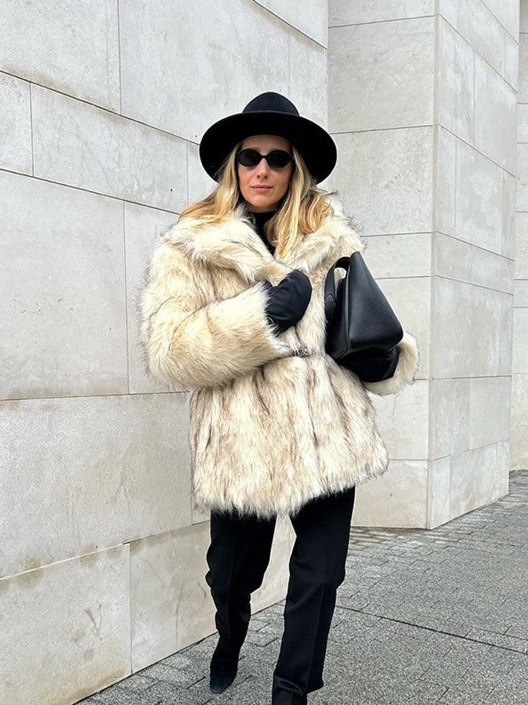Woman in a cream faux fur coat, black hat, and black pants walking outdoors.