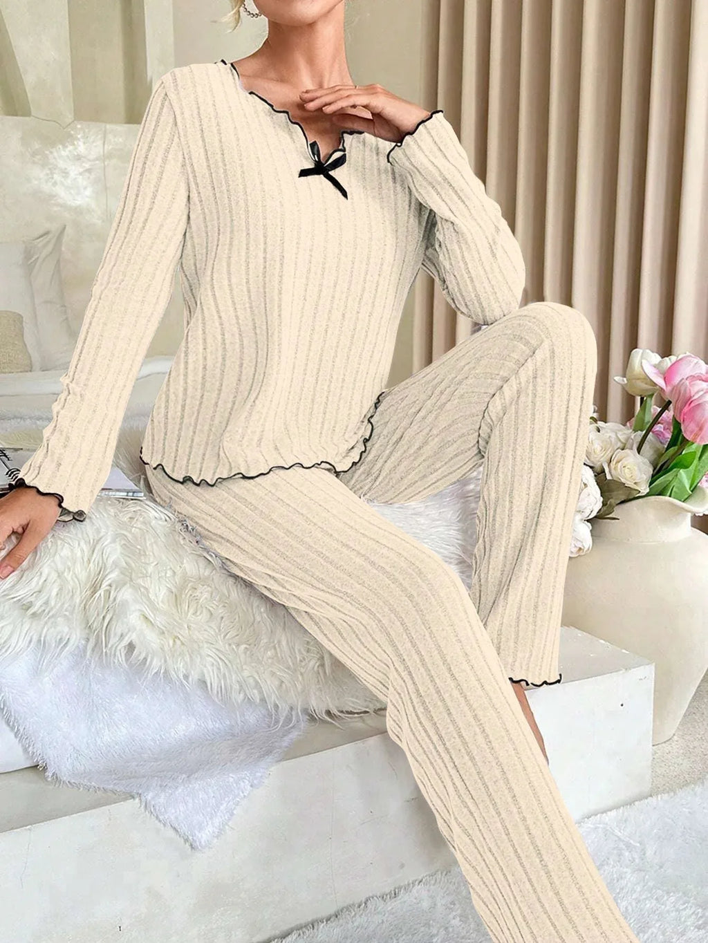 Woman sitting on a bed wearing a cream ribbed knit pajama set with black trim, posing with her hand on her chest.