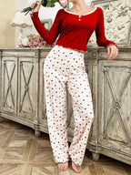 Woman wearing a red long-sleeved top and white pants with a red crown print, leaning against a wooden dresser.