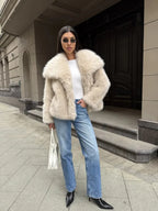 Full-body shot of a woman wearing a cream-colored faux fur jacket, white top, blue jeans, black boots, and sunglasses, holding a white handbag.
