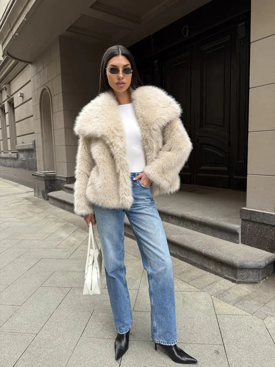 Full-body shot of a woman wearing a cream-colored faux fur jacket, white top, blue jeans, black boots, and sunglasses, holding a white handbag.