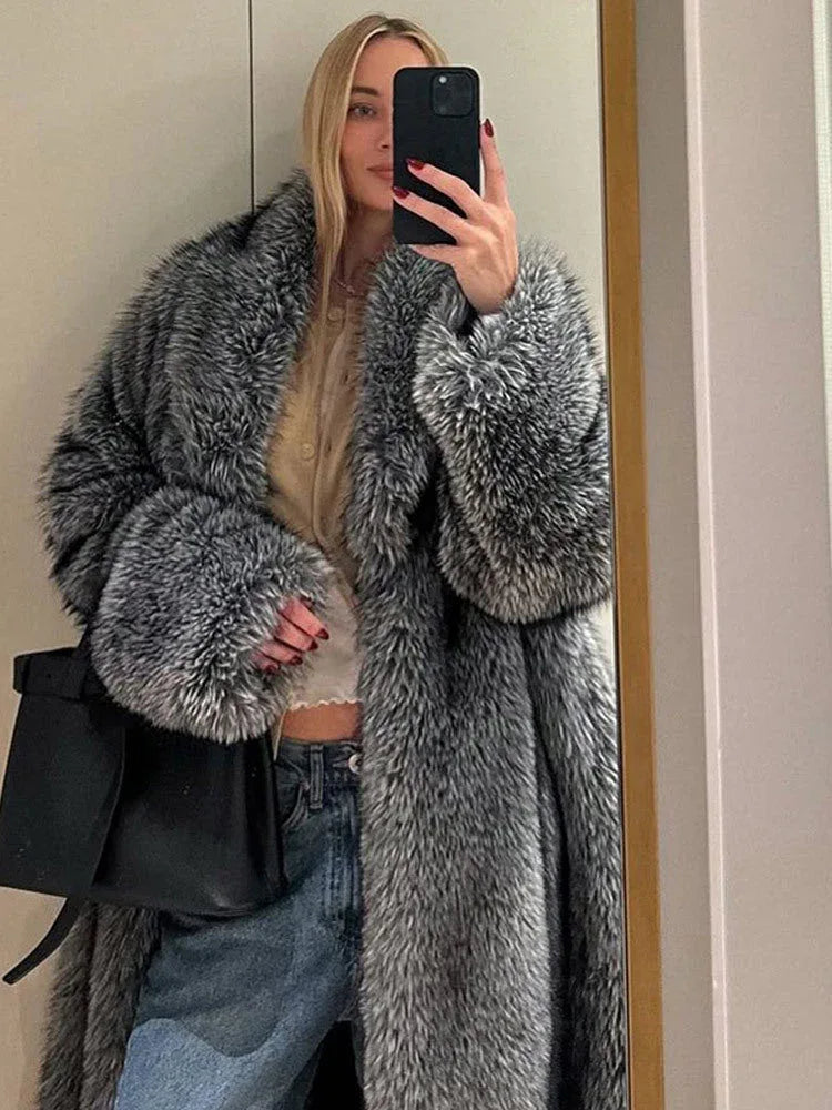 Woman taking a mirror selfie wearing a long grey faux fur coat, a beige top, and blue jeans.