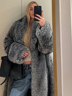 Woman taking a mirror selfie wearing a long grey faux fur coat, a beige top, and blue jeans.