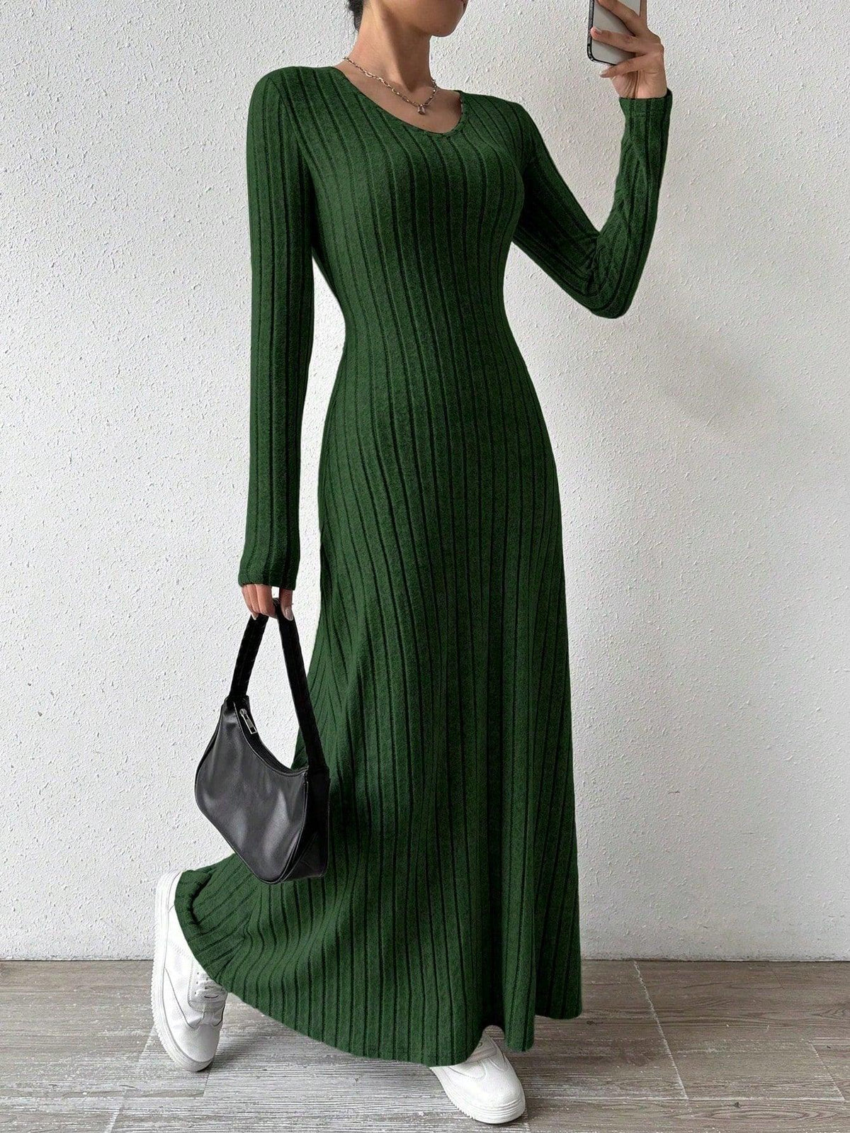 Woman wearing a long-sleeved green ribbed maxi dress, holding a black handbag and wearing white sneakers, posing against a white wall. Main image