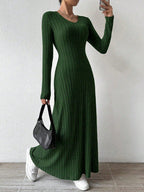 Woman wearing a long-sleeved green ribbed maxi dress, holding a black handbag and wearing white sneakers, posing against a white wall.