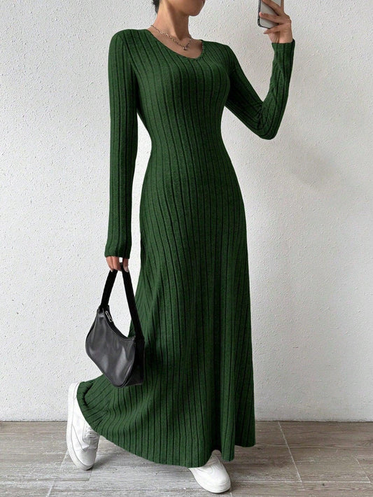 Woman wearing a long-sleeved green ribbed maxi dress, holding a black handbag and wearing white sneakers, posing against a white wall.