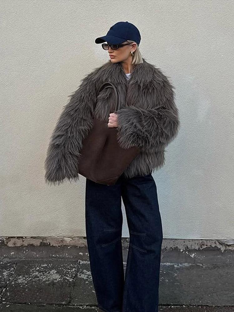 Woman wearing a shaggy grey faux fur jacket, dark wash wide-leg jeans, and a navy baseball cap, standing against a wall.