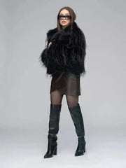 Woman in a black faux fur jacket, brown wrap dress, sheer black tights, and black knee-high boots, posing with hands on hips.