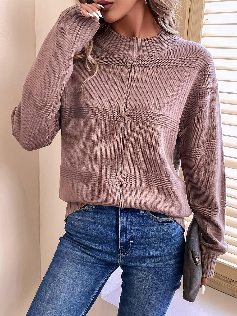 Front view of a woman wearing a mauve knit sweater with a mock neckline and long sleeves, holding a dark clutch.
