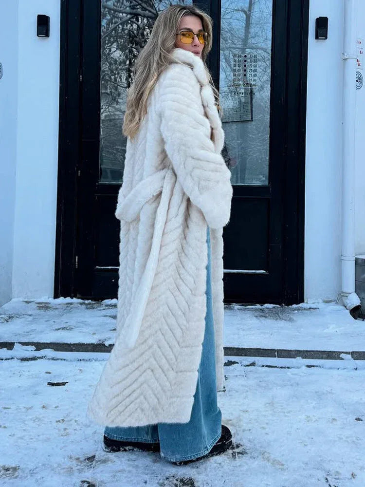 Woman wearing a cream faux fur long coat with a chevron pattern and a belted waist, seen from the back, standing in front of a building with snow.