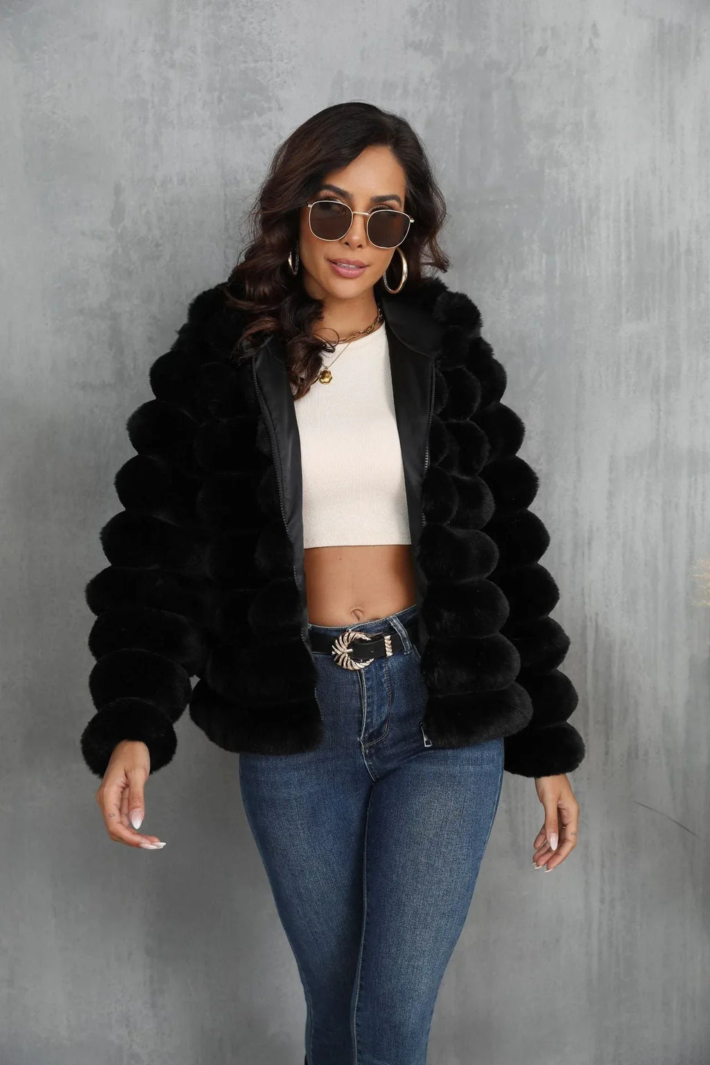 Woman wearing a black faux fur hooded jacket over a white crop top and blue jeans, with sunglasses on.